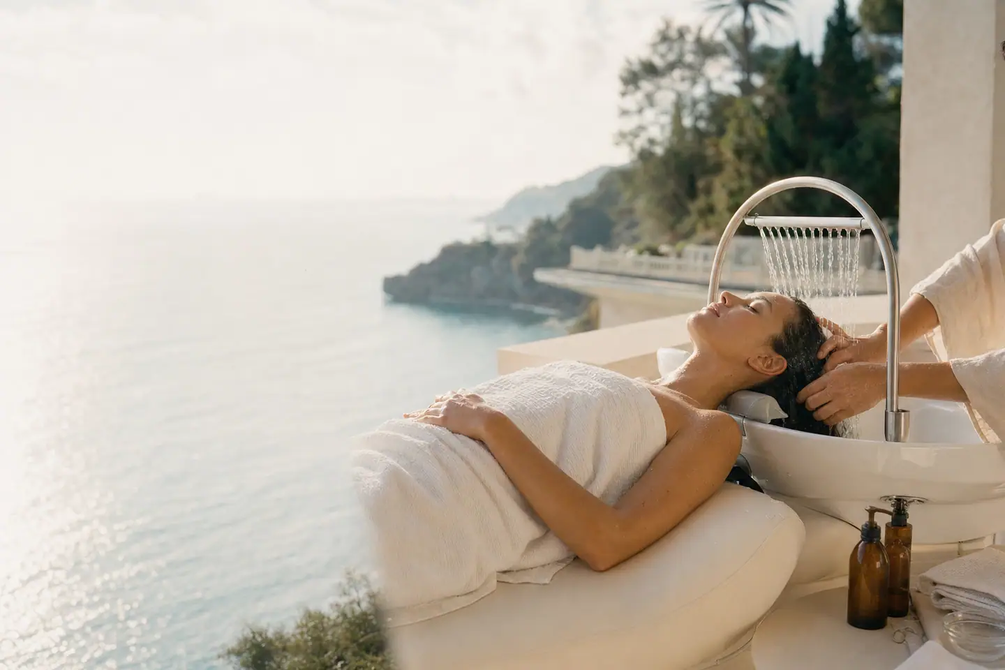 Séance de head spa sur une terrasse face à la mer : allongée sur le lit à vasque, une cliente reçoit un massage du cuir chevelu sous la pluie fine de l'arceau
